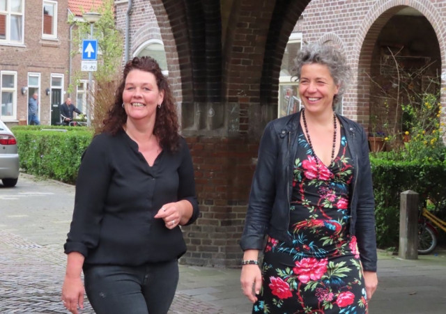 Een foto van twee witte vrouwen. Ze lopen onder een boogje door in de wijk de Hoogte in Groningen. De vrouw links heeft donker, lang krullend haa.r. Ze draagt een zwarte blouse. De vrouw rechts heeft krullend, grijs haar. Ze draagt een jurk met bloemen en een lange kralenketting. Ze lachen allebei.