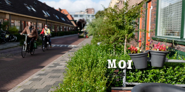 Foto van een straat in een Groningse wijk met op de voorgrond een groene heg met de letters MOI  en twee fietsers op straat