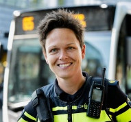 Een foto van een witte vrouw met kort bruin haar. Ze glimlacht en kijkt in de camera. Ze heeft een politieuniform aan en je ziet haar walkie talkie. Op de achtergrond zie je wazig een bus.