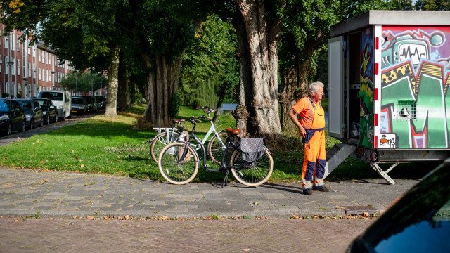 Foto van een man naast twee geparkeerde fietsen in een Groningse wijk met oranje werkkleding aan bij een met graffiti bespoten schaftkeet.  