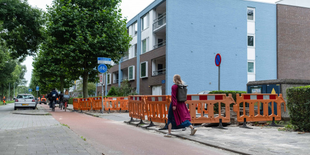 Foto van de Jadestraat in Groningen waar een jongedame in een jurk gehinderd door een opgebroken stoep met oranje hekken ervoor, van de stoep naar het fietspad loopt. In de verte zie je drie fietsers op het fietspad met een de linkerzijde een rij bomen en aan de rechterzijde een lage flat met een lichtblauwe zijgevel.