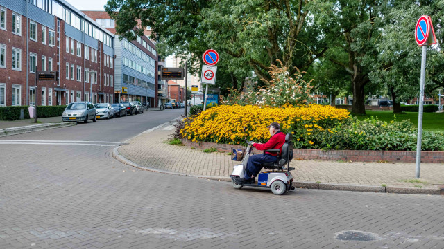 Een foto van een man in een scootmobiel. Hij rijdt over de Zaagmuldersweg in de Oosterpoort. Hij draagt een rode jas en heeft een pet op. Op de achtergrond zie je gele bloemen en een bushalte.