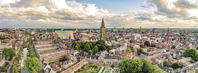 Een luchtfoto van de stad Groningen. Met op de voorgrond veel groen van de Prinsentuin. En daar achter de Martinitoren. Verder is er blauwe lucht met wat wolken.