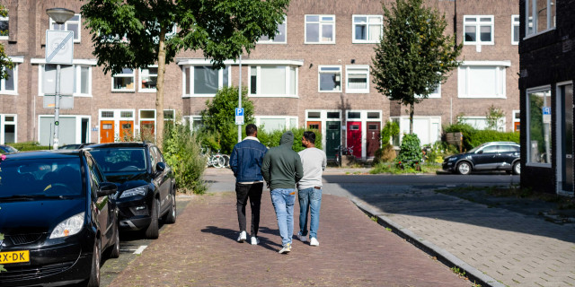 Foto van drie donkere jongemannen lopend op straat in een Groningse wijk. Links staan twee geparkeerde auto's op de achtergrond een rij huizen. 