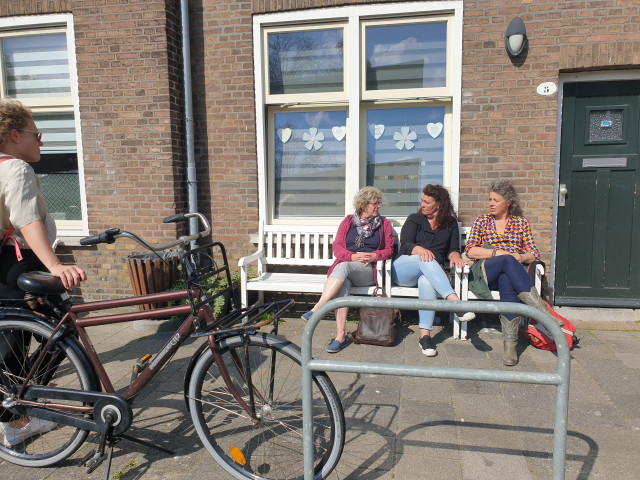 Een foto van een groepje mensen op een bankje in de Groningse wijk De Hoogte. Het bankje staat tegen een voorgevel aan. Op de voorgrond zie je een fiets en een fietsenrekje. De vrouwen hebben alle drie krullend haar.