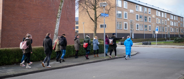 Een groep mensen loopt over de stoep van de Ossehoederstraat in Groningen. We zien ze op de rug. Iemand in een lange blauwe jas loopt over de straat. Iemand vooraan op de stoep wijst naar rechts. Er zijn nieuwe flatgebouwtjes te zien.