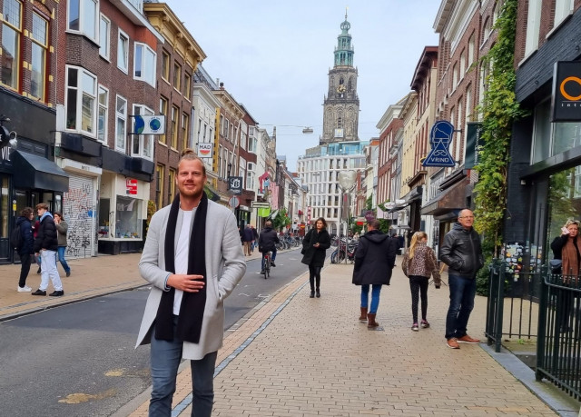 Een foto van een witte man op straat in Groningen. Hij heeft blond haar in een knotje en draagt een lange zwarte sjaal. Hij loopt door de Oosterstraat. Tussen winkelende mensen en winkels.