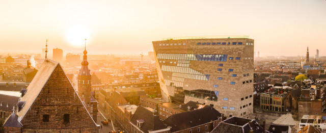 Een luchtfoto van Forum Groningen. Het lijkt een beetje mistig, de zon schijnt fel waardoor de foto een oranje gloed heeft. Je ziet ook de rest van het centrum en bijvoorbeeld het torentje van het Provinciehuis.
