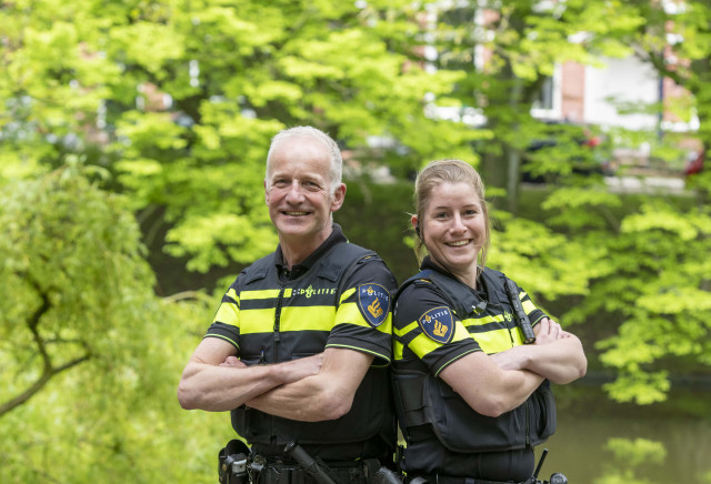 Een foto van twee witte mensen in politieuniform. De ene is een man van middelbare leeftijd.  De ander een jonge vrouw met blond haar. Ze hebben de armen over elkaar en lachen naar de camera. Op de achtergrond is veel groen en een vijver te zien.