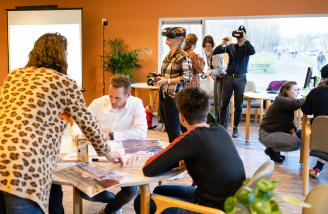Een foto van wutte mensen in een ruimte met tafels en stoelen. Twee mensen dragen een VR-bril. Andere mensen kijken toe en overleggen over foto's en plattegronden.