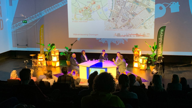 Een foto van een tafelgesprek met witte mannen. Je ziet publiek op de rug. De witte mannen zitten op een podium. Op de achtergrond is een plattegrond van de stad Groningen te zien.